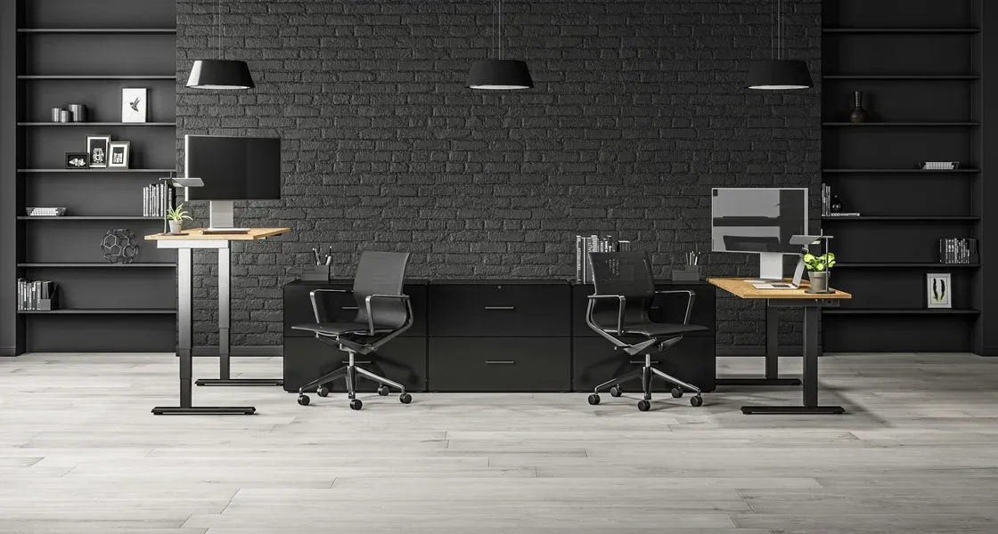 Two standing desks with monitors and chairs against a dark brick wall
